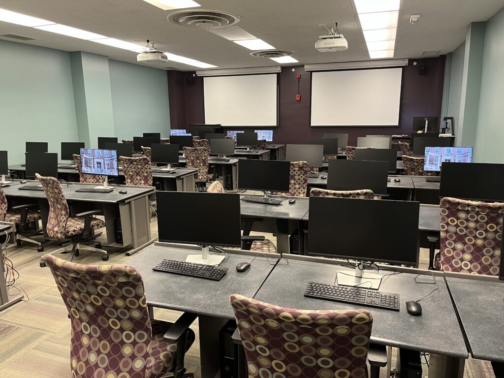 Picture of Wohlers Lab with tables, computers, chairs, and two screens at the front of the room.